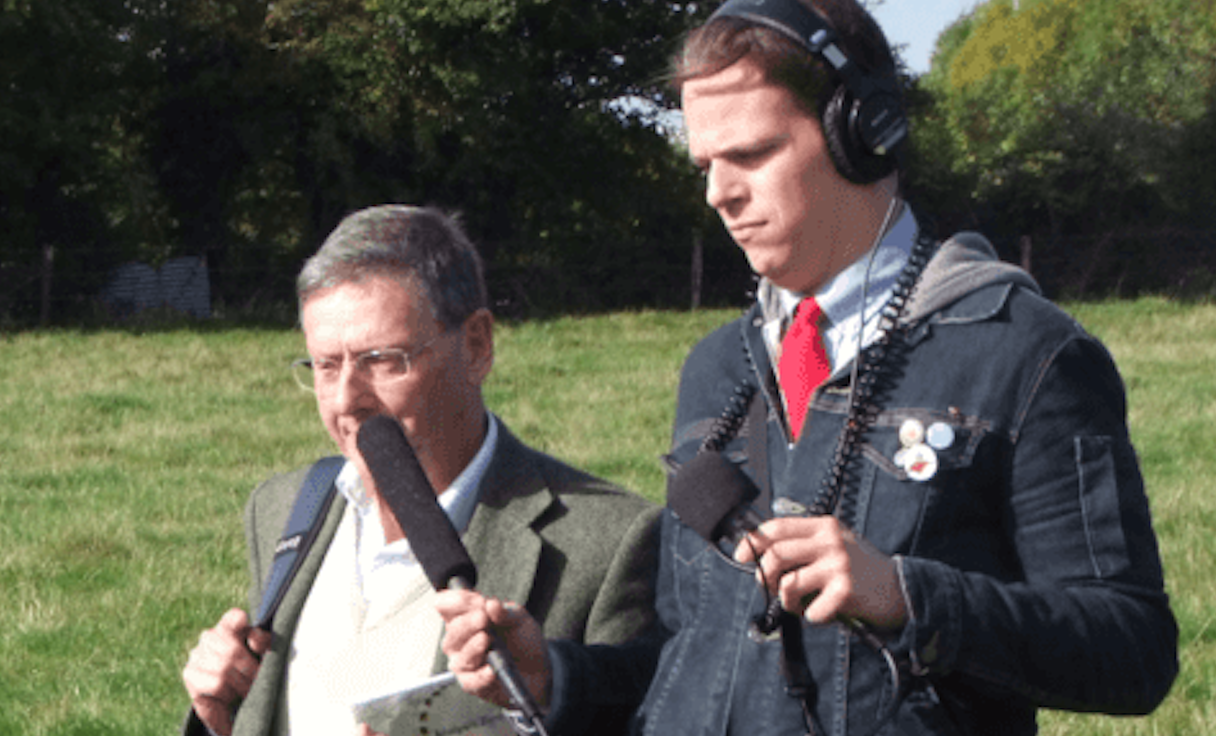 White non-bonary person wearing a jean jacket, blue button down oxford, and red knit tie with headphones one holding a shotgun mic interviewing a white man with a green wool jacket and cream sweater and glasses while walking across a grass field with trees in the background