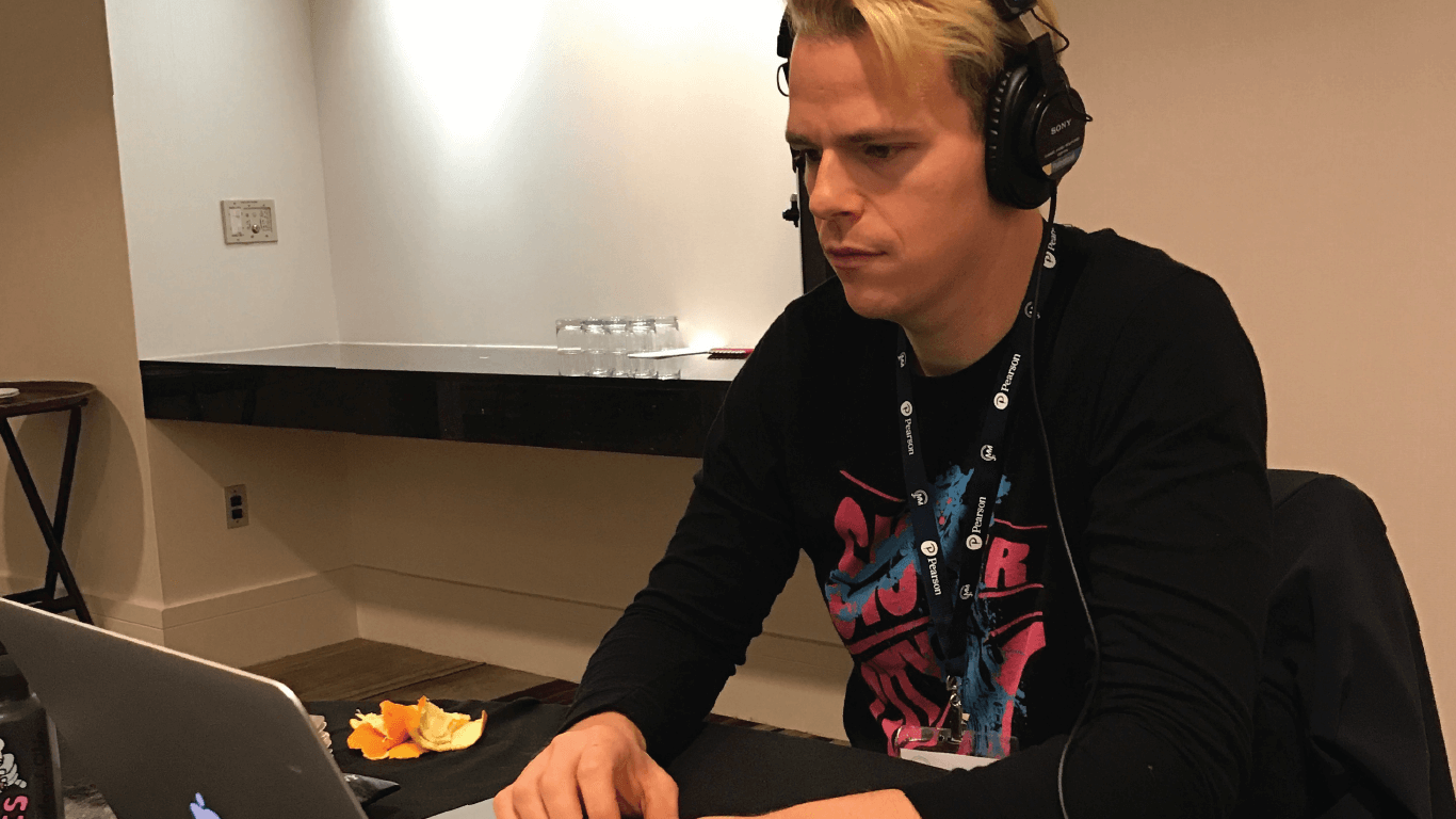 image of a white non-binary person with swept to the side bleached hair with headphones on at a laptop with a whiteboard behind them