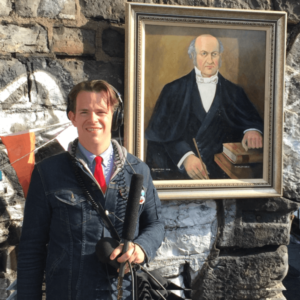 White non-bonary person wearing a jean jacket, blue button down oxford, and red knit tie with headphones one holding a shotgun mic standing in front of a sand stone brick wall with a portrait of an old white man from the 1800s (William Rowan Hamilton)